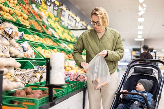 Casualy dressed mother buying fruits and vegetables in supermarket grocery store with her infant baby boy child in stroller
