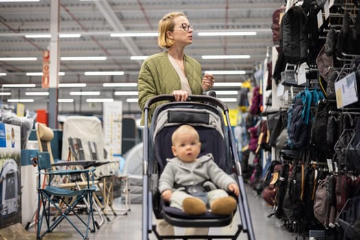 Casualy dressed mother choosing sporty shoes and clothes products in sports department of supermarket store with her infant baby boy child in stroller