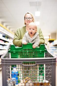 Mother pushing shopping cart with her infant baby boy child down department aisle in supermarket grocery store. Shopping with kids concept