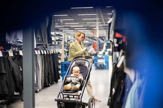 Casualy dressed mother choosing sporty shoes and clothes products in sports department of supermarket store with her infant baby boy child in stroller