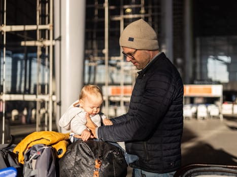 Fatherat comforting his tired infant baby boy child sitting on top of luggage cart in front of airport terminal station while traveling wih family