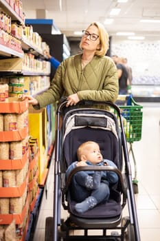 Casualy dressed mother choosing canned products in department of supermarket grocery store with her infant baby boy child in stroller