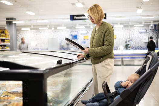 Casualy dressed mother choosing frozen products in department of supermarket grocery store with her infant baby boy child in stroller