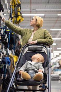 Casualy dressed mother choosing sporty shoes and clothes products in sports department of supermarket store with her infant baby boy child in stroller