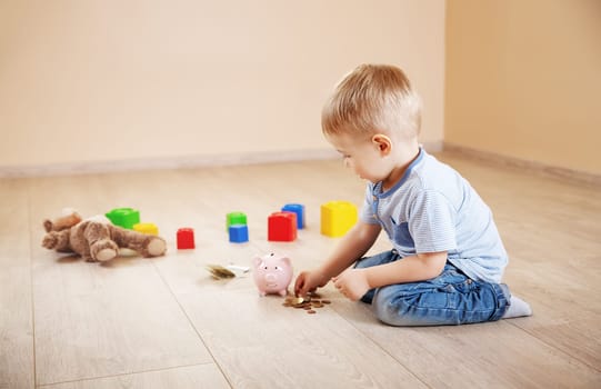 Cute boy sitting on the floor indoor and inserting euro coins in piggybank. Concept of the economy and family budget.