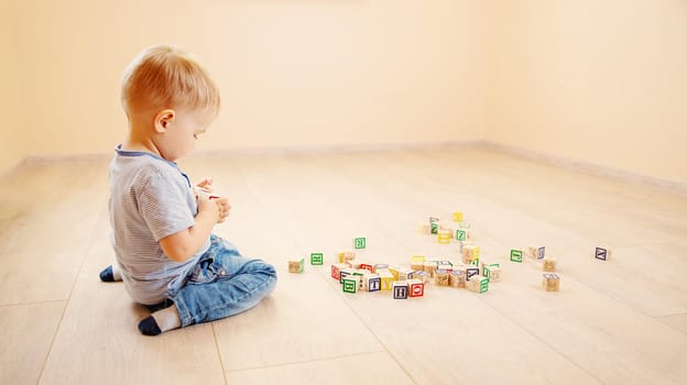 Little boy sitting on the floor indoor and playing with wooden cubes. Concept of preschool and kindergarden.