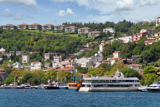 View from the sea of the green mountains of the Europian side of Bosphorus strait, with docked boats, traditional houses and dense trees in a summer day