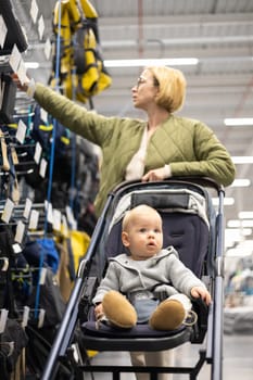 Casualy dressed mother choosing sporty shoes and clothes products in sports department of supermarket store with her infant baby boy child in stroller