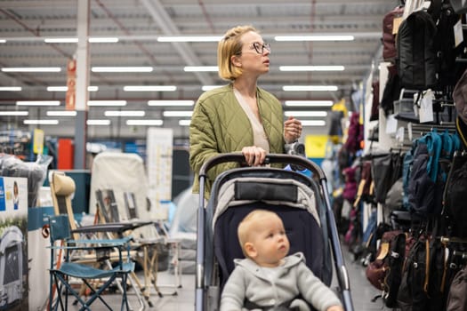 Casualy dressed mother choosing sporty shoes and clothes products in sports department of supermarket store with her infant baby boy child in stroller