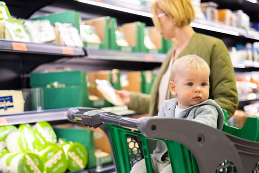 Casualy dressed mother choosing products in department of supermarket grocery store with her infant baby boy child in shopping cart
