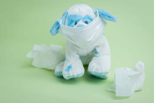 A white and blue stuffed dog wearing a surgical mask surrounded by used tissues representing the idiomatic phrase "sick as a dog."""