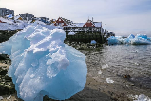 Big piece of blue ice laying among the stones on the shore with modern Inuit building on the hill at the fjord, Nuuk city, Greenland