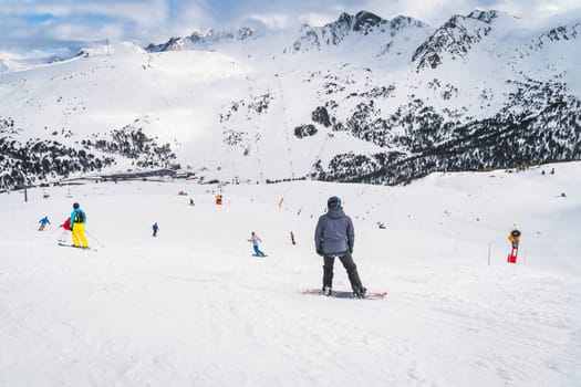 Snowboarders and skiers riding down the mountain towards valley below with snow capped mountain peaks in background, Andorra, Pyrenees