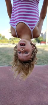 Crazy girl hangs upside down on the sports ground. Exercises on sports equipment. A Caucasian child of 8 years old with blond hair in a striped dress. Fun girl surprised and opened her mouth.