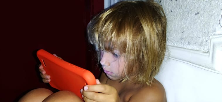 Cute little girl plays with a tablet computer. A child of 7 years old with a serious face and blond hair looks into a gadget. Copy space. Girl in the room. Dark and light wall. At home in the evening.