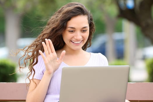 Happy woman using laptop in a video call sitting on a bench in a park