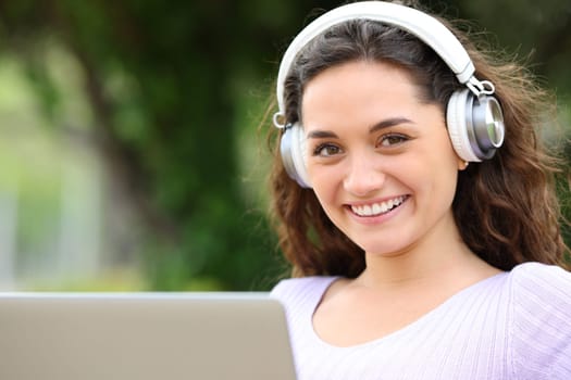 Happy woman using laptop and headset looks at you in a park