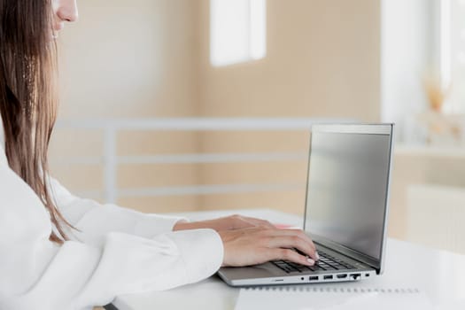 Woman hands typing on the laptop with empty blank white screen mockup. Computer is laying on the white table. Blurred background. Person working from home concept. Copy space area for text