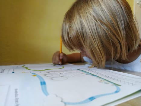 A 6-7 year old girl is reading a book or textbook and smiling. The child has blond hair, dark skin and glasses on his face. The child sits at the desk. yellow wall in the background.