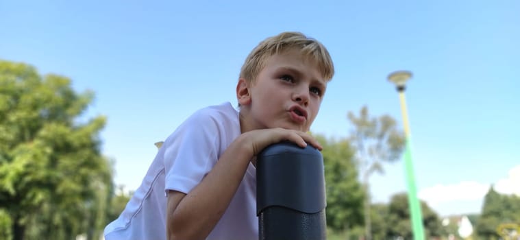 Pensive boy on the playground. The child lingered on top of the metal structure, and says something. Boy with blond hair.