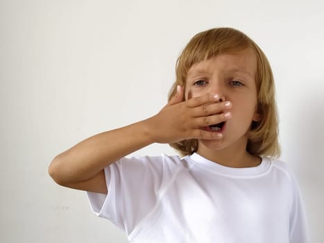 A little cute girl 6-7 years old expresses emotional joy and a smile on a white background. A child with tanned skin. White T-shirt. Copy space. The child yawns, covering his mouth with his hand.