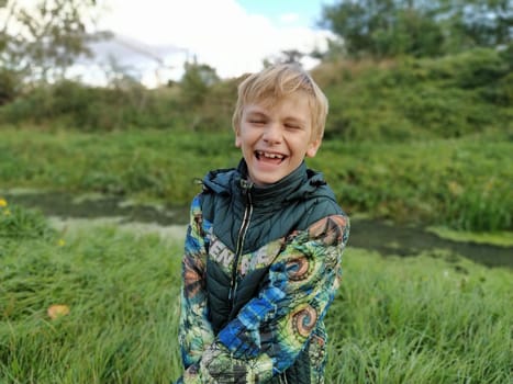 A blond boy, 8 years old, laughs in the fall on a walk by a swampy pond. A violent emotion of fun on the face of a child. Everyday warm clothes. Green jacket