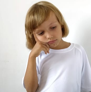 Little disappointed girl 6-7 years old with blond hair, white background. Tanned skin. White T-shirt. Copy space. The girl raised her right hand and put her index finger to her temple. Hairstyle Bob