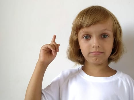 A little cute girl 6-7 years old with blond hair on a white background, pointing up. A child with tanned skin. White T-shirt. Copy space. Girl with her hand points to something. Bob hairstyle.