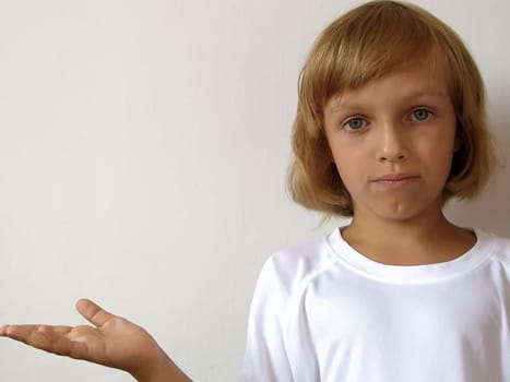 A little cute girl 6-7 years old with blond hair expresses emotional joy and a smile on a white background. A child with tanned skin. White T-shirt. Copy space. Girl with her hand points to something.