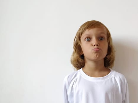 Little cute girl with blond hair on a white background. Tanned skin. White T-shirt. Copy space. The girl rolled her eyes and pursed lips. Facial expressions, surprise. Keeping a secret. Hairstyle Bob