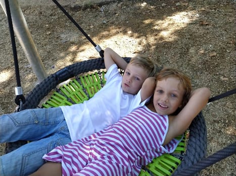 Children swing on a spider-web swing. Boy and girl, brother and sister in the summer on the playground. Swinging the swing. Boy 8 years old in a white Tshirt. Girl 6 years old in a striped pink dress