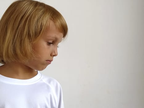 Girl on a white background. The child looks down and to the side. Copy space. White T-shirt. Caucasian blond hair, dark tanned skin. Head side. Bob hairstyle.