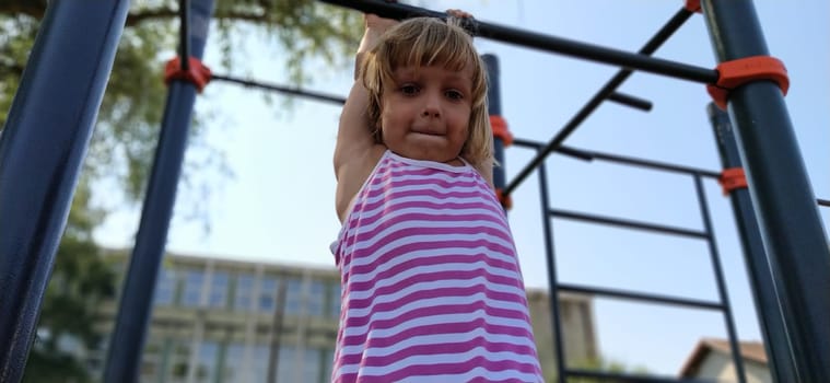 A child in a striped dress hangs on a metal bar and tries to pull up on his arms. Sports or playground. Outdoor exercise.