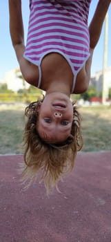 Girl upside down. A child with blond hair in a striped pink dress hangs from a bar in the playground. The hair hangs down, the mouth is half open. Girl looking at the camera