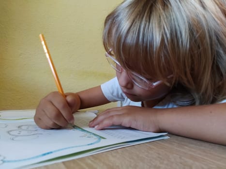 A 6 or 7 year old girl draws in a book. The child does his homework. Blond hair and pink-rimmed glasses. Training and education concept.