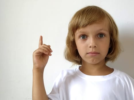 A little cute girl 6-7 years old with blond hair on a white background, pointing up. A child with tanned skin. White T-shirt. Copy space. Girl with her hand points to something. Bob hairstyle.