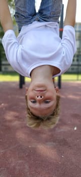 The boy hangs upside down on the sports ground. Exercises on sports equipment. Caucasian child 8 years old with blond hair. Sports and fun