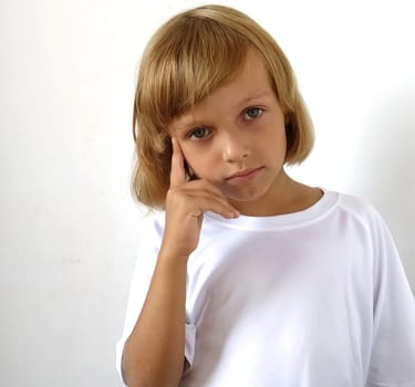 Little cute girl 6-7 years old with blond hair on a white background. Tanned skin. White T-shirt. Copy space. The girl raised her right hand and put her index finger to her temple. Hairstyle Bob