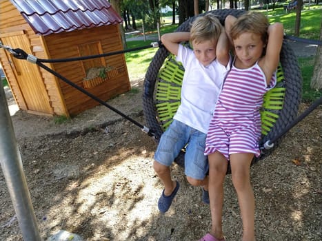 Children swing on a spider-web swing. Boy and girl, brother and sister in the summer on the playground. Swinging the swing. Boy 8 years old in a white T-shirt. Girl 6 years old in a striped pink dress.