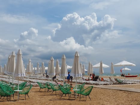 Anapa, Russia, August 15, 2021 Sun loungers and umbrellas on the sandy sea beach after the rain. Beach season. Beach cafe with wicker chairs. Tourists walking people. A storm warning. Cumulus clouds.