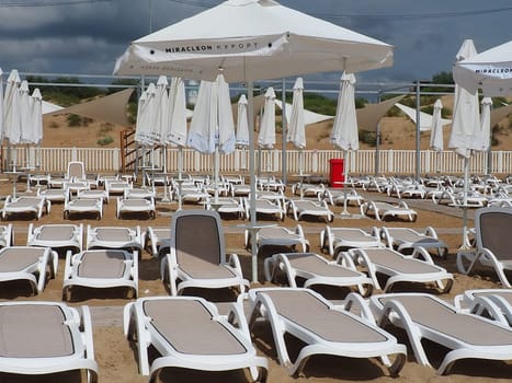 Anapa, Russia, August 15, 2021 Wet sunbeds and umbrellas on the sea beach during heavy rain storm. Rolled up parasol on the beach against dark cloudy sky. Invasion of a typhoon, cyclone or storm.