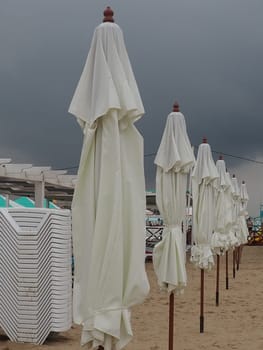 Wet sunbeds and umbrellas on the sea beach during heavy rain storm. Rolled up parasol on the beach against dark cloudy sky. Invasion of a typhoon, cyclone or storm on the sea or ocean. Beach season.