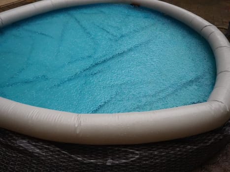 Rubber round pool filled with water. Blue water in the pool. Large drops of rain fall on the surface of the water. Rainy weather at the hotel. No one in the pool due to bad weather.