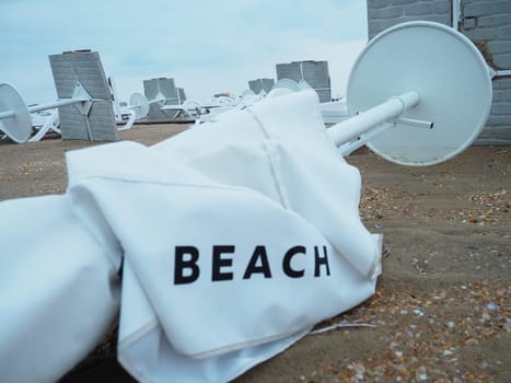 A beach umbrella lay folded on a sandy beach after bad weather. Storm at sea. End of the beach season due to typhoon and cyclone. Loungers, umbrellas and hotel furniture. Beach word written on white.