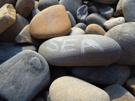Oval stones, large smooth sea pebbles, illuminated by the summer sun. The word sea is written on the stone. Summer vacation in the resort, wellness, sea adventures and pleasures. Stone gray background