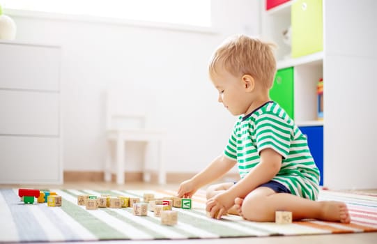 Cute boy sitting on the carpet indoor and playing with wooden cubes. Concept of preschool and kindergarden.