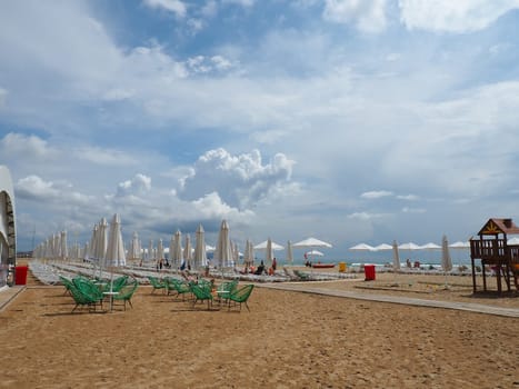 Anapa, Russia, August 15, 2021 Sun loungers and umbrellas on the sandy sea beach after the rain. Beach season. Beach cafe with wicker chairs. Tourists walking people. A storm warning. Cumulus clouds.