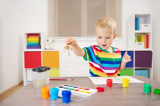 Cute boy drawing picture by using watercolors and couache indoors. Concept of leisure and kindergarten.