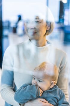 Thoughtful young mother looking trough window holding his infant baby boy child while waiting to board an airplane at airport terminal departure gates. Travel with baby concept