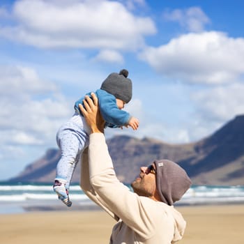 Father enjoying pure nature holding and playing with his infant baby boy sun in on windy sandy beach of Famara, Lanzarote island, Spain. Family travel and parenting concept
